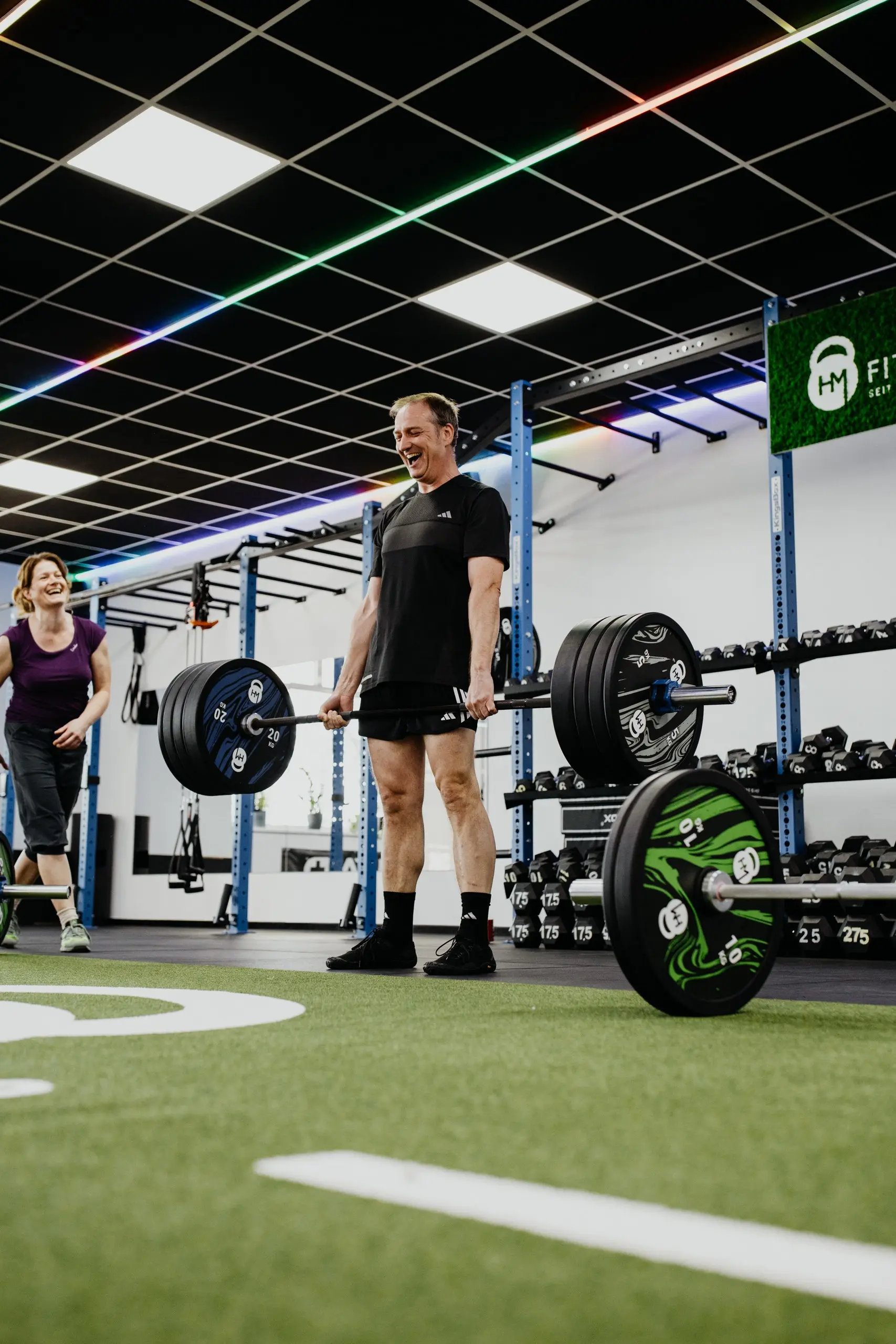 Mitglied steht mit der beladenen Langhantel aufrecht, im Hintergrund lacht eine Trainingspartnerin